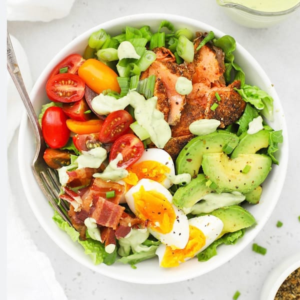 A vibrant salmon salad with avocado, cherry tomatoes, bacon, and hard-boiled eggs, drizzled with dressing.