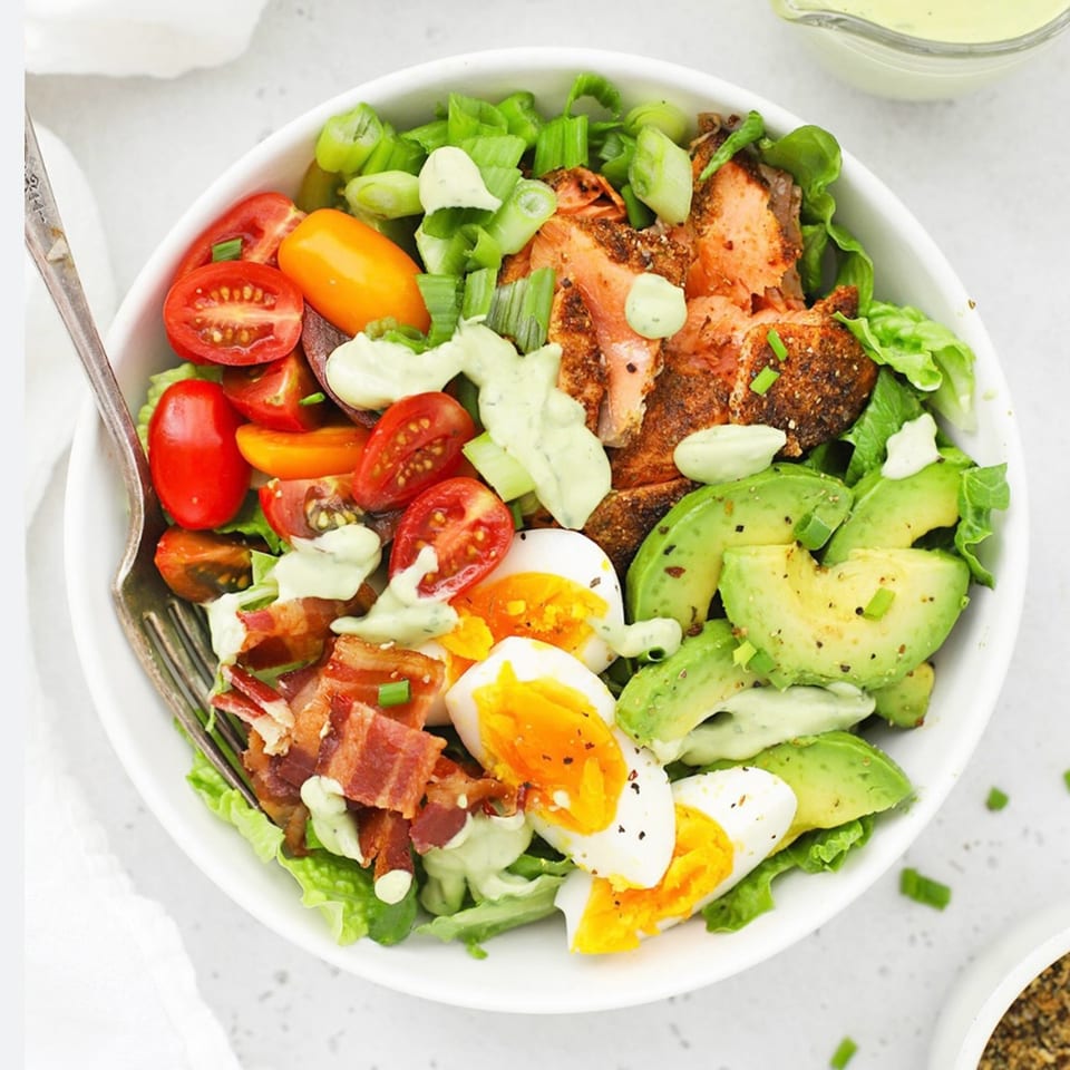 A vibrant salmon salad with avocado, cherry tomatoes, bacon, and hard-boiled eggs, drizzled with dressing.