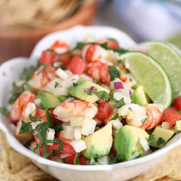A white bowl of shrimp ceviche with avocado, tomato, onion, and cilantro, garnished with lime wedges.