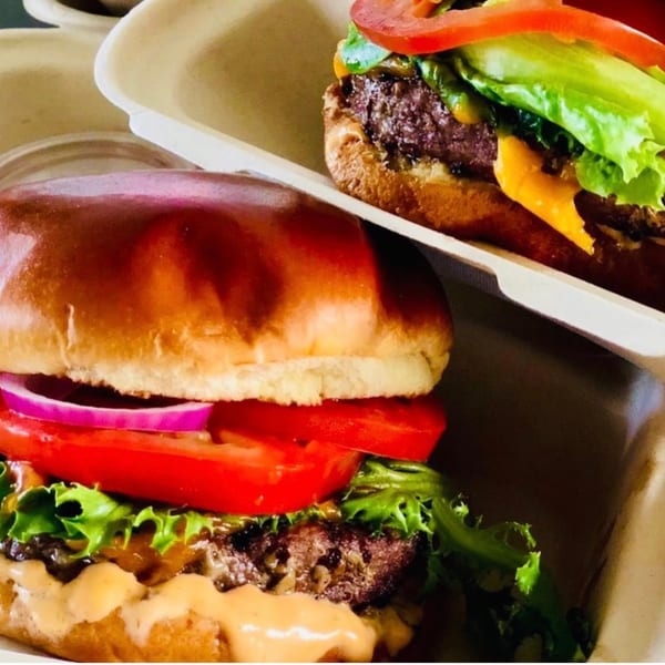Two juicy burgers with lettuce, tomato, onion, and sauce in take-out containers from The Cabana Bar & Grill.