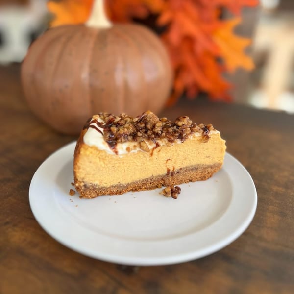 A slice of pumpkin cheesecake with whipped cream, nuts, and chocolate drizzle on a white plate.